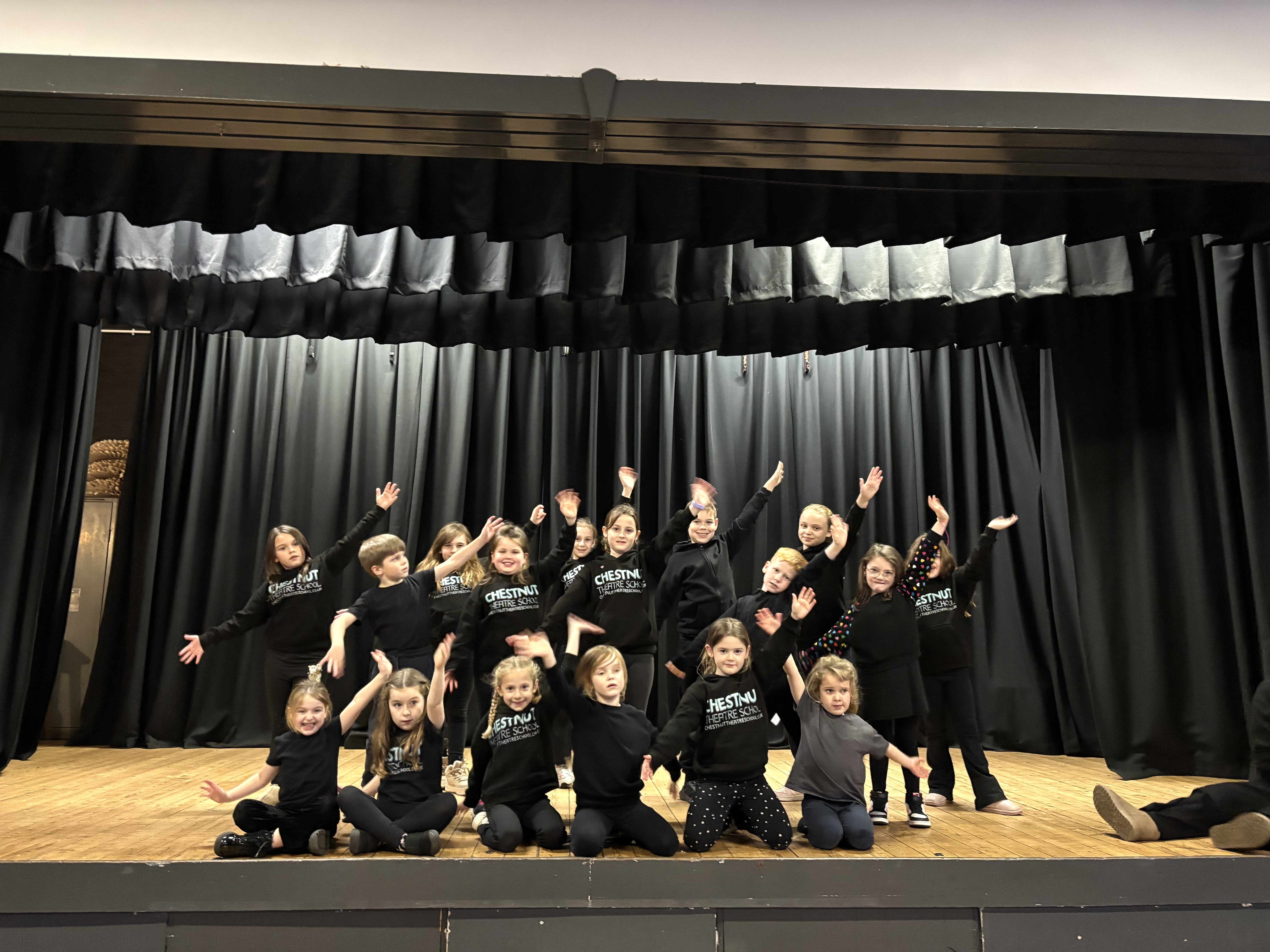 A large group of children in black posing on stage with their arms stretched up and out in a dance move